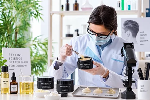 Scientist wearing gloves and a lab coat, formulating hair pomade in a laboratory setting with testing trays and hair care products displayed.